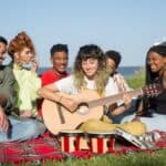 多元文化團隊在海邊野餐，欣賞吉他演奏與音樂交流，展現國際勞工的合作與友誼。.