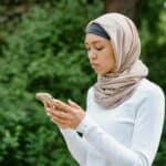 手機使用中的年輕穆斯林女性，戶外自然背景，展現多元文化融入的職場與生活情境。.