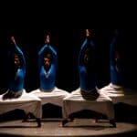 深色背景下表演的演員，穿著藍色上衣與白色寬褲，展現傳統舞蹈姿勢。.